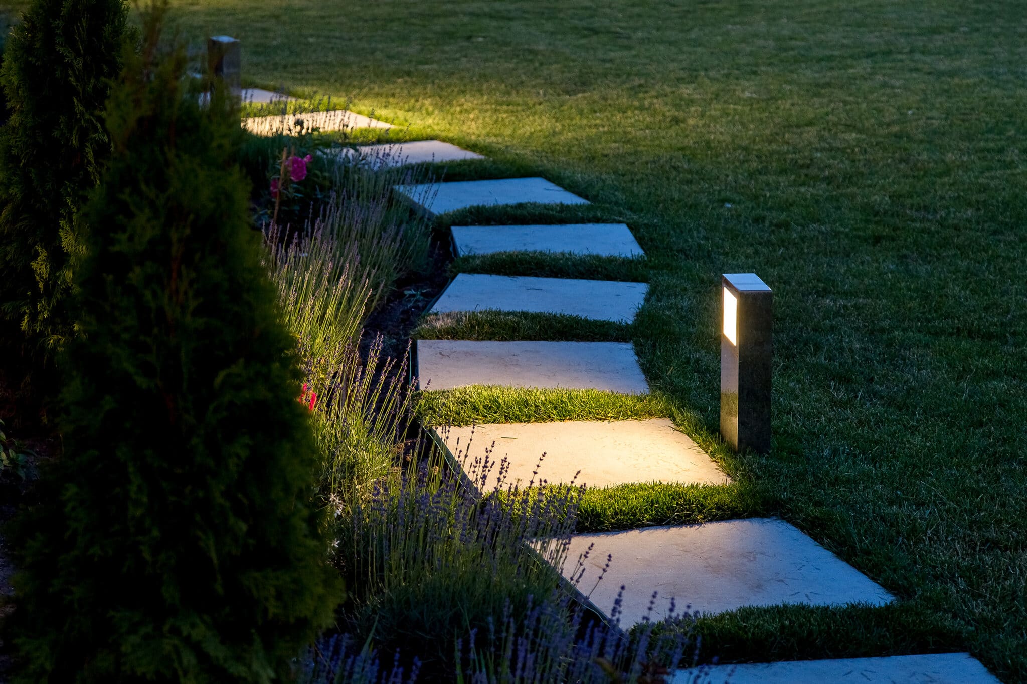 A marble path of square tiles is illuminated by landscape lighting, casting a warm glow in a backyard garden.